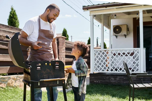 Feliz homem americano africano no avental cozinhar milho na churrasqueira e olhando para o filho no quintal — Fotografia de Stock