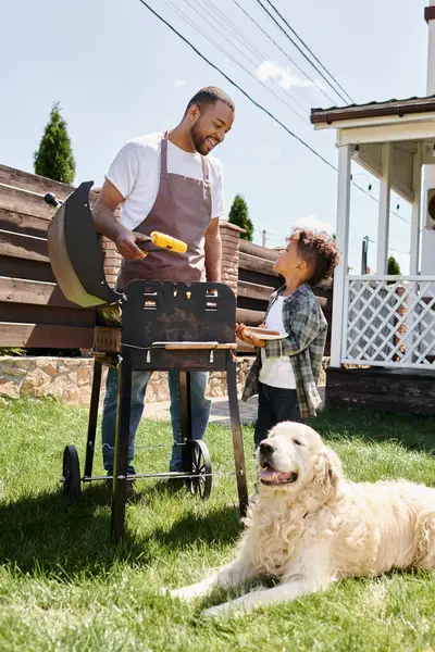 Cheerful african american man in apron cooking corn on bbq grill and looking at son on backyard — Stock Photo