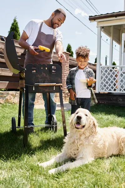 Positive african american man in apron cooking corn on bbq grill and looking at dog near son — Stock Photo