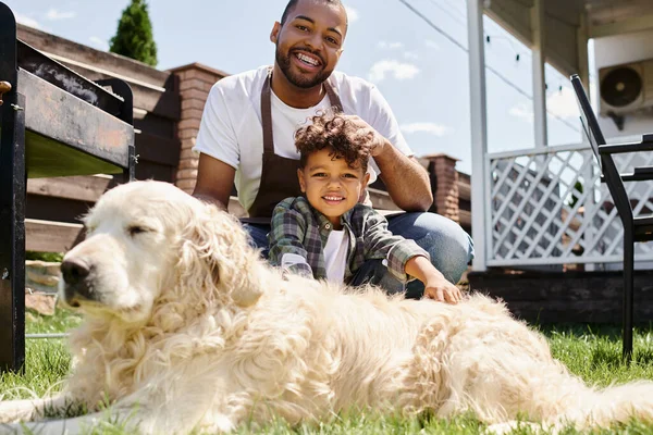 Felice uomo africano americano in grembiule toccando i capelli ricci del figlio e seduto vicino al cane di famiglia all'aperto — Foto stock