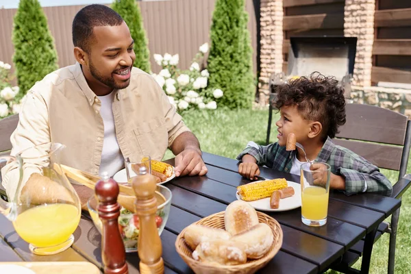 Feliz pai americano africano almoçando com filho encaracolado ao ar livre, salsichas grelhadas e milho — Fotografia de Stock