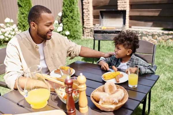 Alegre afroamericano padre comiendo almuerzo con rizado hijo al aire libre, salchichas a la parrilla y maíz - foto de stock