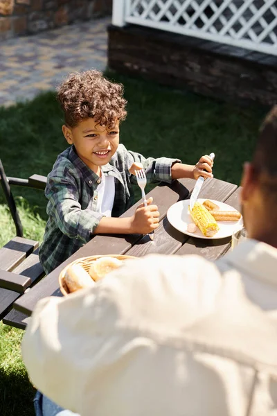 Menino americano africano encaracolado feliz comendo salsichas e milho grelhado enquanto olha para o pai ao ar livre — Fotografia de Stock