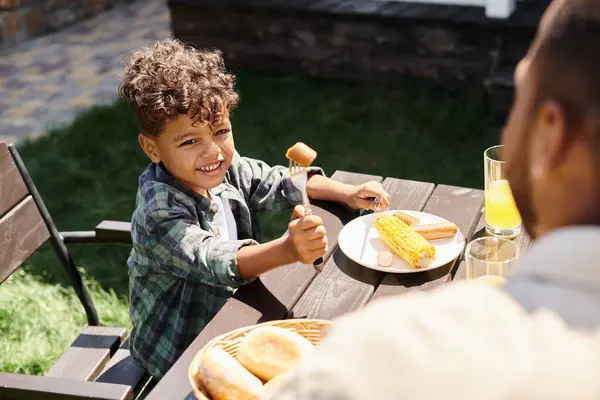 Alegre rizado afroamericano chico comiendo salchichas y maíz a la parrilla mientras mira a padre al aire libre - foto de stock