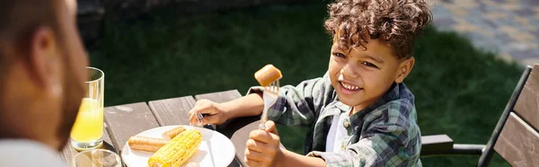 Gioioso riccio africano americano ragazzo mangiare salsicce e mais alla griglia mentre guardando il padre, banner — Foto stock