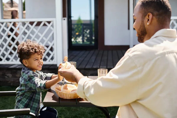 Щасливий афроамериканський батько, проходячи свіжу булочку, щоб кучерявий усміхнений син, маючи сімейний барбекю — стокове фото