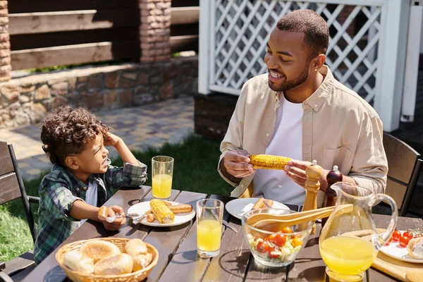 Felice padre afroamericano avendo famiglia bbq con riccio figlio sul cortile di casa, mais alla griglia — Foto stock