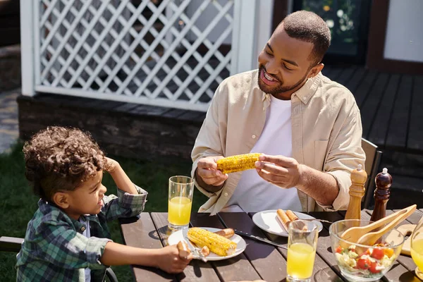 Gioioso padre afroamericano avendo famiglia bbq con riccio figlio sul cortile di casa, mais alla griglia — Foto stock