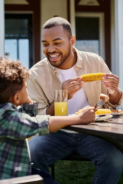 Positivo afroamericano padre tener familia barbacoa con rizado hijo en el patio trasero de la casa, parrilla de maíz - foto de stock