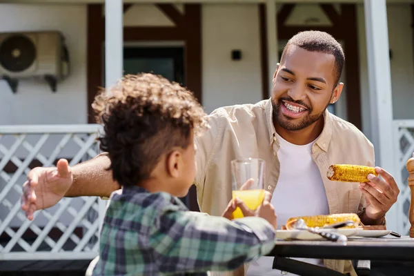 Hombre afroamericano positivo que tiene barbacoa familiar con hijo rizado en el patio trasero de la casa, maíz a la parrilla - foto de stock