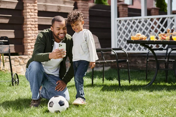 Happy african american father and son looking at smartphone and standing near football on backyard — Stock Photo