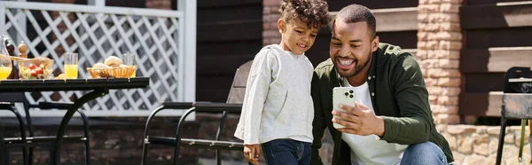 Happy african american father and son looking at smartphone and standing on backyard, banner — Stock Photo