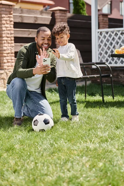 Happy african american father and son looking at smartphone during video call  on backyard — Stock Photo