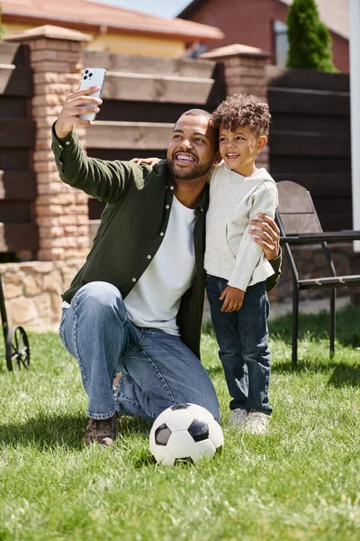 Feliz afroamericano padre e hijo mirando el teléfono inteligente mientras toma selfie en el patio trasero - foto de stock
