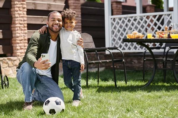 Happy african american father and son holding smartphone while standing on backyard of house — Stock Photo