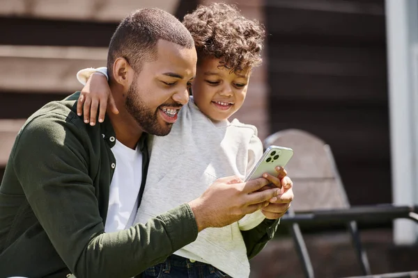 Homem americano africano alegre em aparelho e filho usando smartphone no quintal da casa nos subúrbios — Fotografia de Stock
