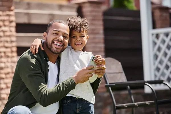 Hombre afroamericano positivo en frenos e hijo usando teléfono inteligente en el patio trasero de la casa en los suburbios - foto de stock