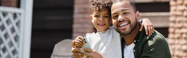 Positive african american man in braces and son using smartphone on backyard of house, banner — Stock Photo