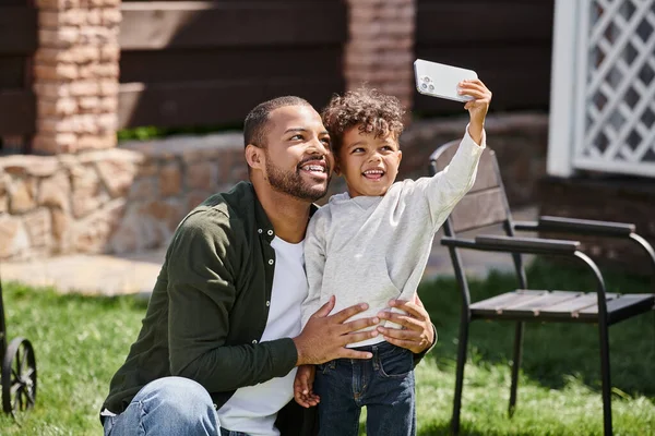 Niño afroamericano feliz tomando selfie en teléfono inteligente con su padre en aparatos ortopédicos en el patio trasero - foto de stock