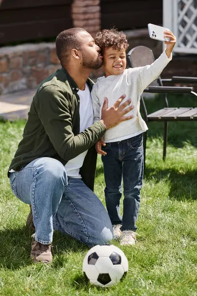 Lindo africano americano niño tomando selfie en smartphone con su padre en patio trasero de la casa, beso - foto de stock