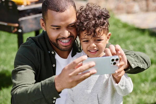 Щасливий афроамериканський хлопчик бере селфі на смартфон зі своїм батьком у дужках на задньому дворі — стокове фото