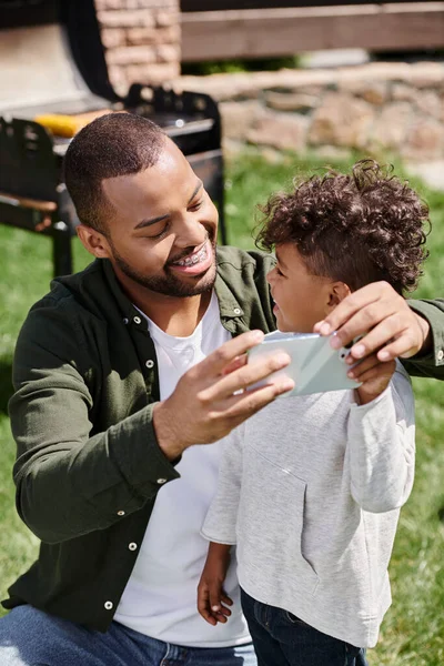 Happy african american father in braces taking selfie on smartphone with his son on backyard — Stock Photo