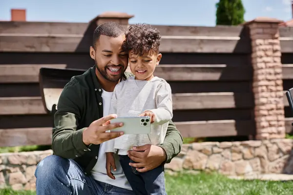 Gioioso padre afroamericano in bretelle scattare selfie su smartphone con suo figlio in cortile — Foto stock