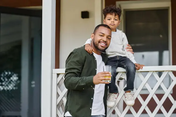 Freudiger afrikanisch-amerikanischer Vater in Hosenträgern, der Orangensaft hält und seinen Sohn auf der Veranda umarmt — Stockfoto