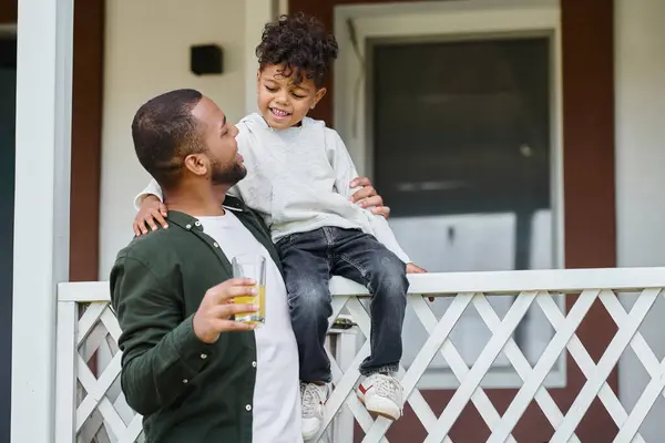 Alegre afroamericano padre en aparatos ortopédicos celebración de jugo de naranja y abrazar a su hijo sentado en porche - foto de stock