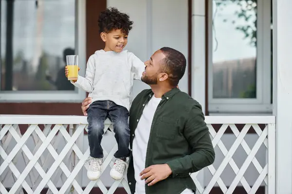 Freudiger afrikanisch-amerikanischer Mann in Zahnspange, der Orangensaft hält und seinen Sohn auf der Veranda umarmt — Stockfoto