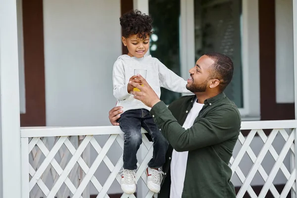 Glücklich afrikanisch-amerikanischer Vater in Zahnspange hält Orangensaft und umarmt niedlichen Sohn sitzt auf der Veranda — Stockfoto