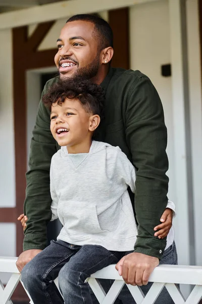 Joyeux garçon afro-américain assis sur le porche et embrassant son père dans des bretelles sur la cour arrière de la maison — Photo de stock