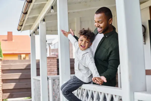 Happy moments, african american boy sitting on porch and holding hands with joyful father in braces — Stock Photo