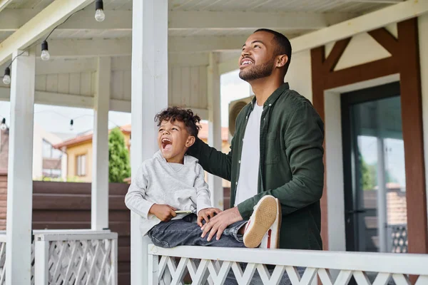 Excited african american father and son smiling and sitting on porch of house, family portrait — Stock Photo