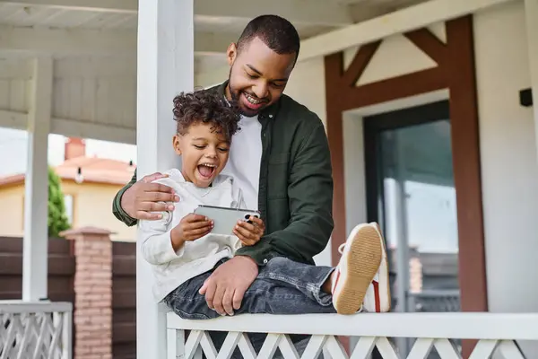 Freudig afrikanisch-amerikanischer Vater umarmt aufgeregten Sohn auf Veranda sitzend und Smartphone in der Hand — Stockfoto