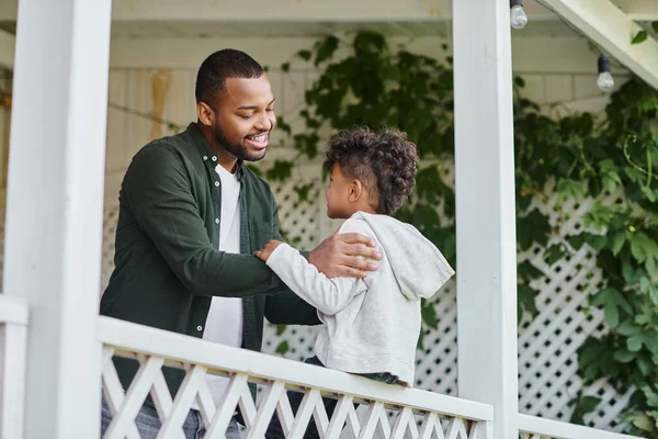 Happy african american father in braces hugging cute curly son sitting on porch on house in suburbs — Stock Photo