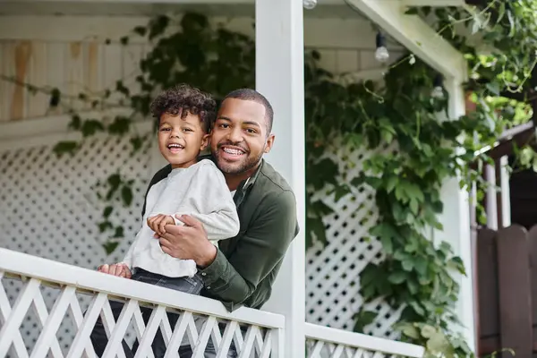 Happy african american father in braces hugging cute curly son and sitting on porch on house — Stock Photo