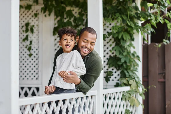 Felice uomo africano americano in bretelle ridere e abbracciare il figlio riccio mentre seduto sul portico — Foto stock