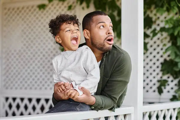 Astonished african american father and son looking away surprisingly while sitting near white fence — Stock Photo