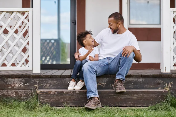 Щасливий афроамериканський чоловік в брекетах обіймає кучерявого сина, сидячи на ганку сучасного будинку — стокове фото
