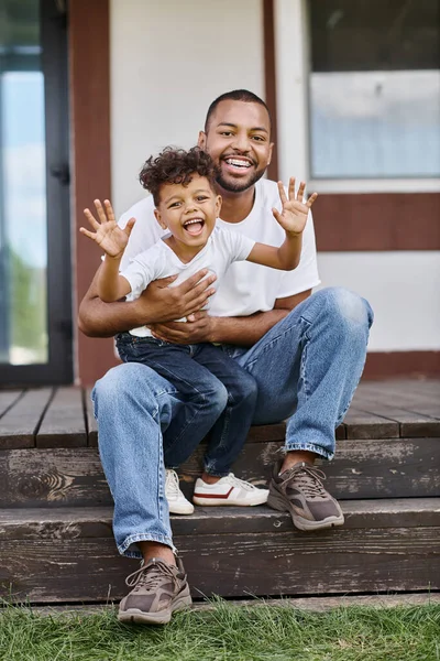 Веселий афроамериканський чоловік в брекетах обіймає збудженого сина, сидячи на ганку сучасного будинку — стокове фото