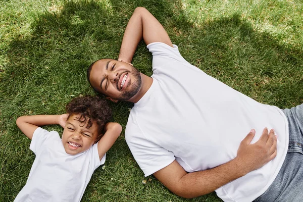 Top view of cheerful african american father and son lying on green lawn on backyard, family photo — Stock Photo