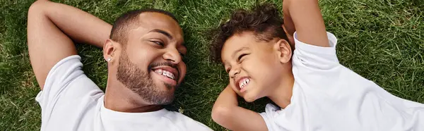 Top view of cheerful african american father and son lying on green lawn on backyard, family banner — Stock Photo