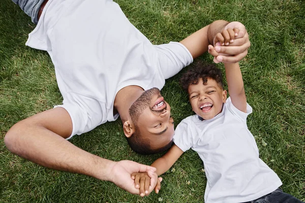 Top view of happy african american father and son lying on lawn on backyard and holding hands — Stock Photo