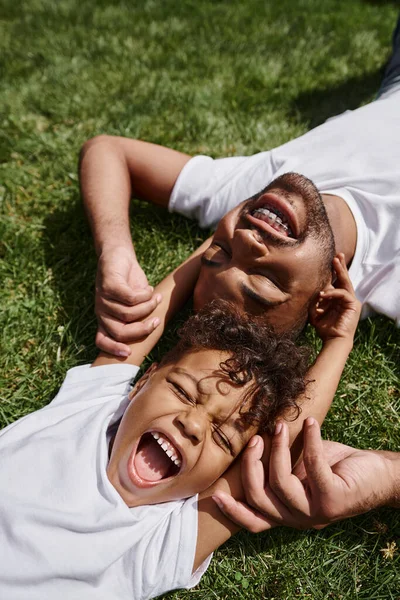 Top view of joyful african american father and son lying on green grass on backyard, family photo — Stock Photo