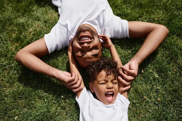 Top view of cheerful african american father and son lying on green lawn on backyard, family fun — Stock Photo