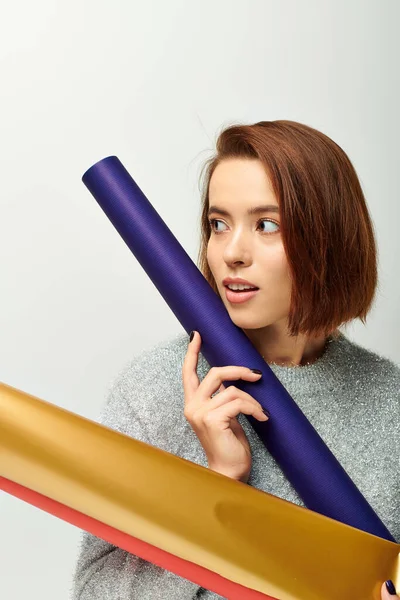 Pretty young woman with short hair holding colorful wrapping paper on grey backdrop, Christmas time — Stock Photo