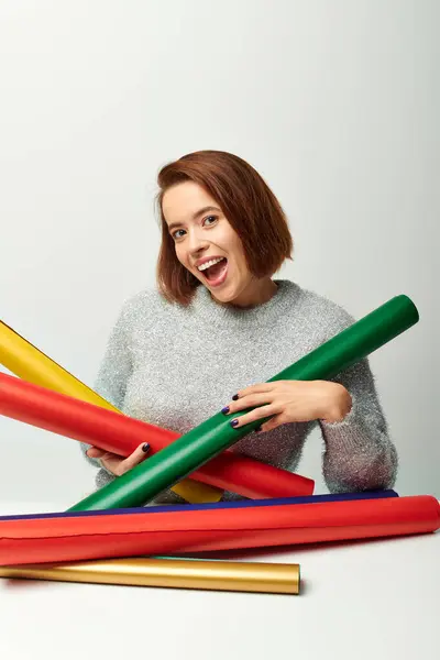 Joyful woman in sweater holding colorful wrapping paper on table with grey wall, Christmas time — Stock Photo