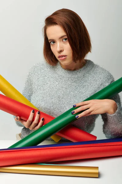 Brunette woman in sweater holding colorful wrapping paper on table with grey wall, Christmas time — Stock Photo