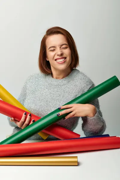 Season of joy, happy woman in sweater holding Christmas gift paper on table with grey backdrop — Stock Photo